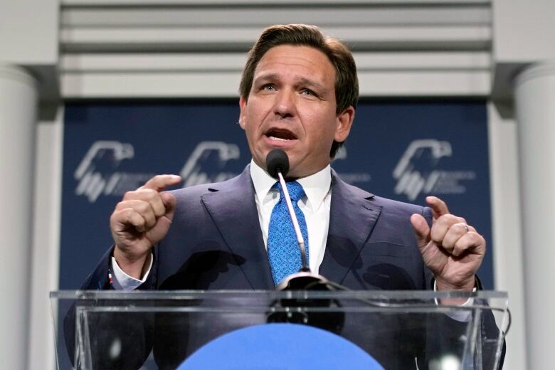 A man in a suit stands at a microphone podium, raising his hands and pointing with his index fingers. 