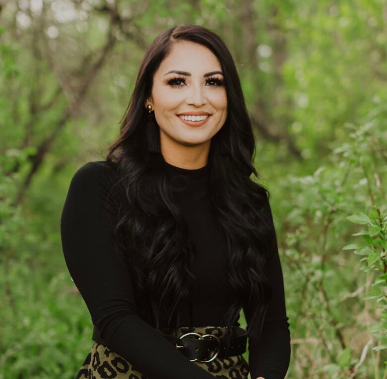 Woman with long dark hair stands in a forest and smiles at the camera wearing a black turtleneck. 