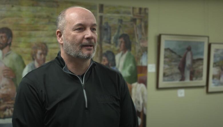 A man wearing a black sweater stands in front of a piece of artwork.