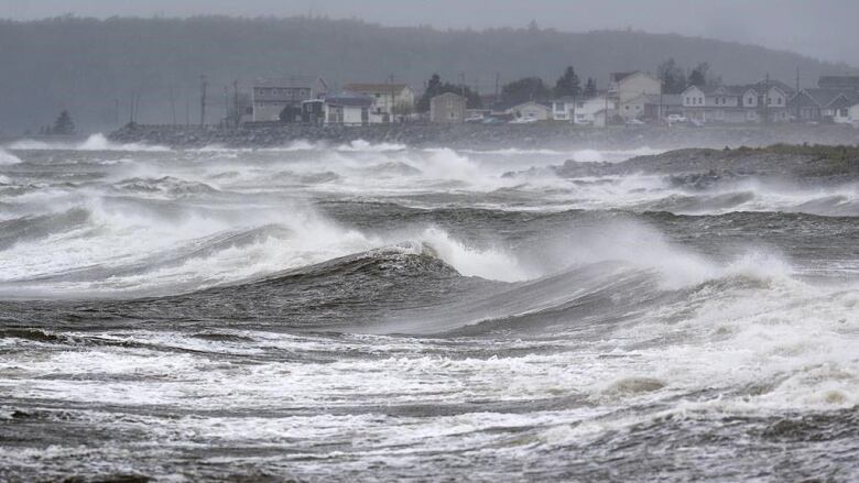 Scene of violent waves with a settlement in the background. There is mist in the air.