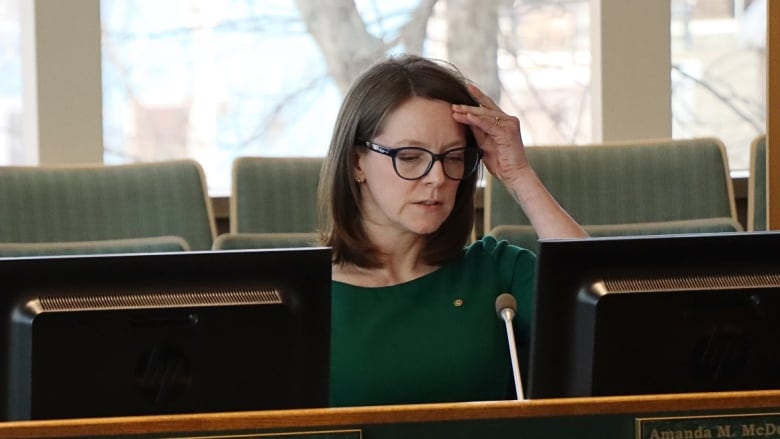 A woman with shoulder-length hair and glasses wearing a green top sits behind a desk.