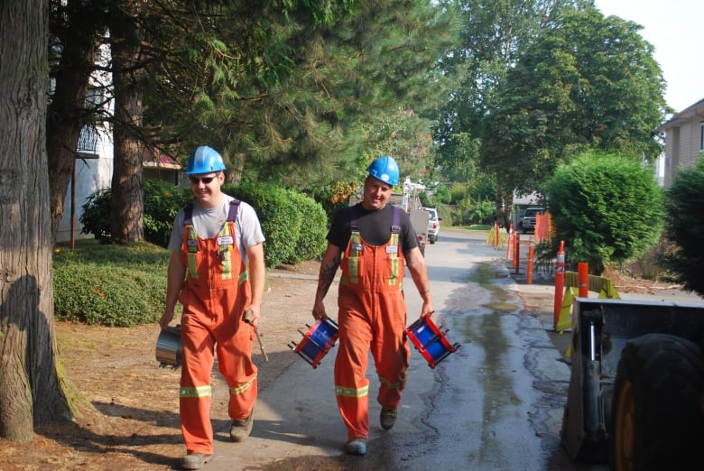 Two men in orange coveralls, blue hard hats and t-shirts carry equipment along a sidewalk.