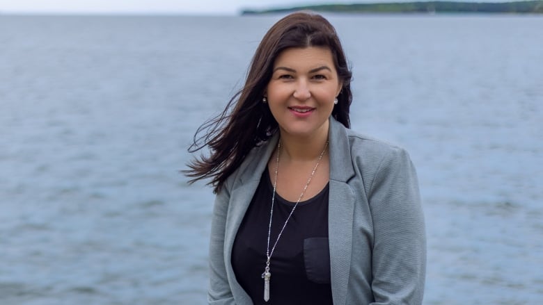 A dark haired woman sits on a rock with the ocean behind her. She is looking at the camera.