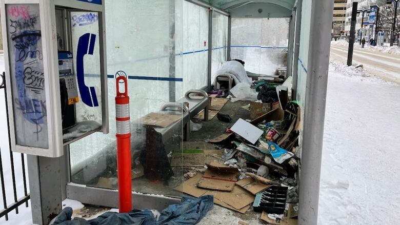 A view into a glass bus shelter shows articles of clothing and other items scattered everywhere. In the foreground, just outside the shelter, is a payphone covered in graffiti.