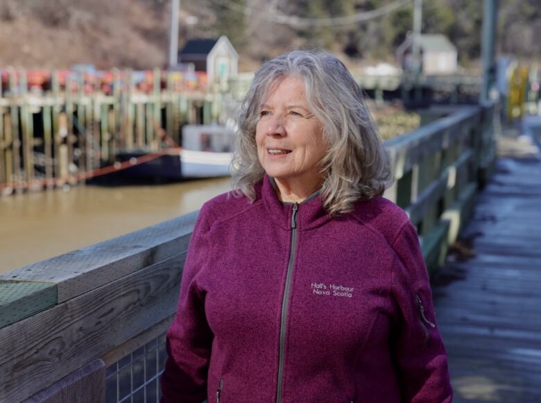 A woman in a purple jacket stands on a walkway with a fishing boat visible in the water behind her.