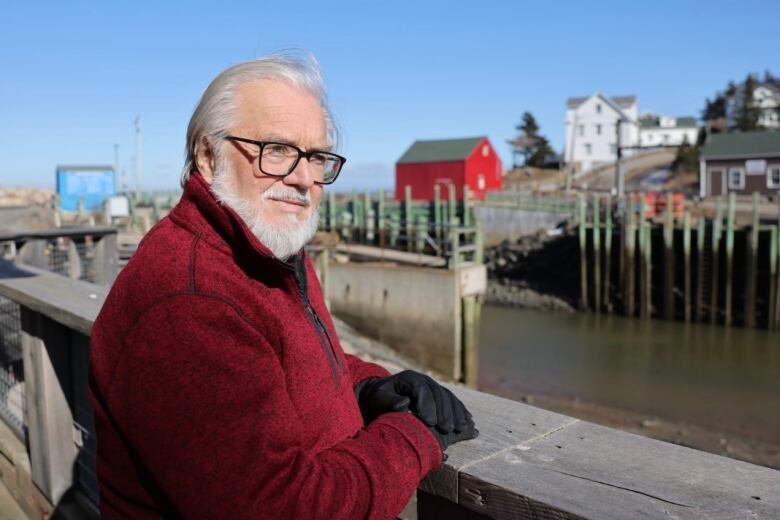 A man in a red jacket stands by the harbour with a few buildings in the background.