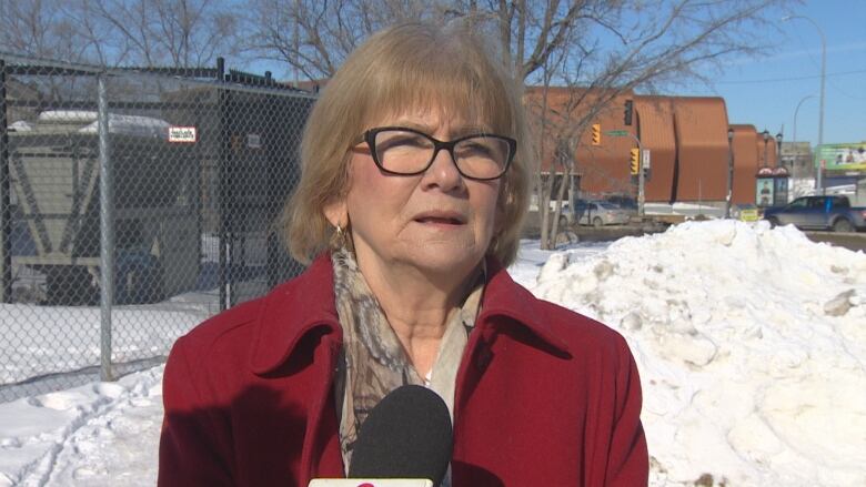 A woman in black glasses and bobbed blonde hair, speaks to a camera. She is outside in the winter and wearing a red coat.