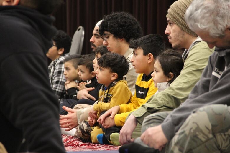 People seated on prayer mats, including children, listen to the speaker.