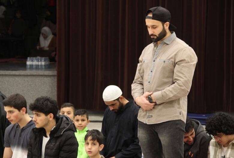 A man, standing on his feet, bows his head in prayer, while other people are either seated or on their knees.