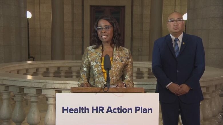 A Black woman in a yellow top and beige coloured jacket stands behind a podium that says 