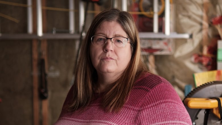 A woman wearing a pink and white striped sweater stands in front of a table of a tools in a garage. She looks frustrated. 