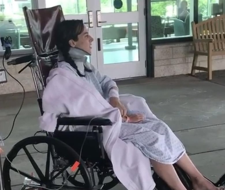 A young woman sits in her a wheel chair outside a hospital while wearing a neck brace.