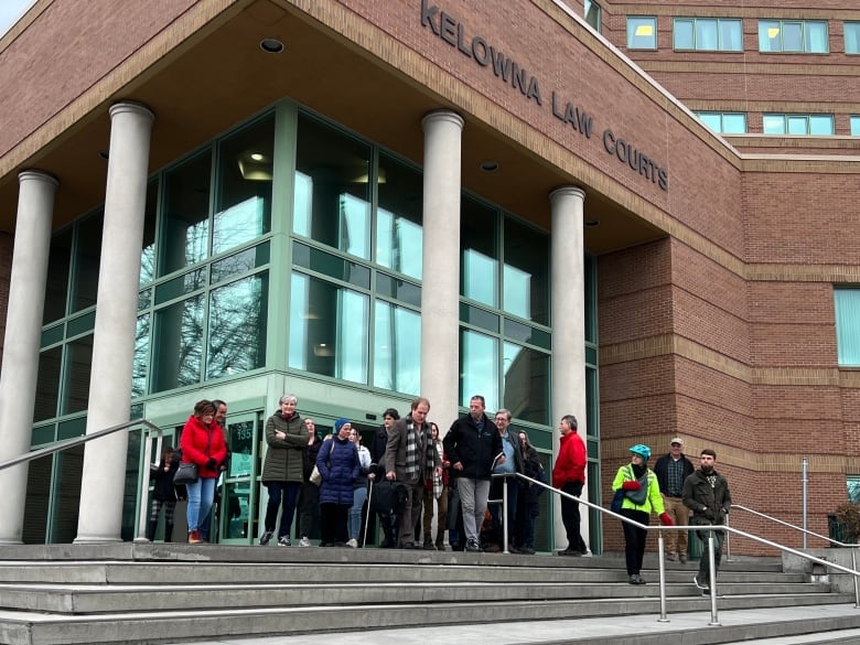 A man in a suit jacket and pants carrying a briefcase is seen leaving the Kelowna courthouse with about 20-30 supporters following him.