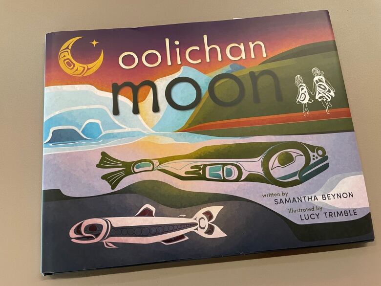 A children's book titled Oolichan Moon shows a yellow moon, a Nisga'a inspired drawing of an oolichan fish and two young girls walking and holding hands.