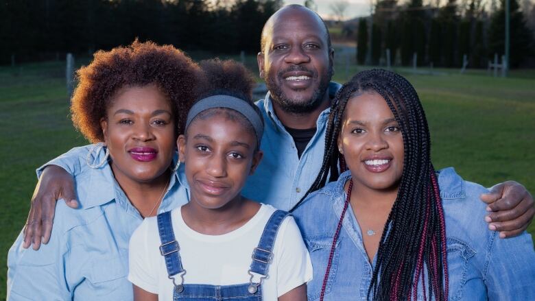 A family of four huddle together and pose for a family portrait. A Black woman with short curly hair wearing a denim button up shirt stands to the left, with her husband next to her, a taller Black man also in a denim button up shirt. A young Black woman stands centered in front of them with a white tee shirt and overalls, and next to her is another young Black woman with her hair in long braids and a denim button up shirt.