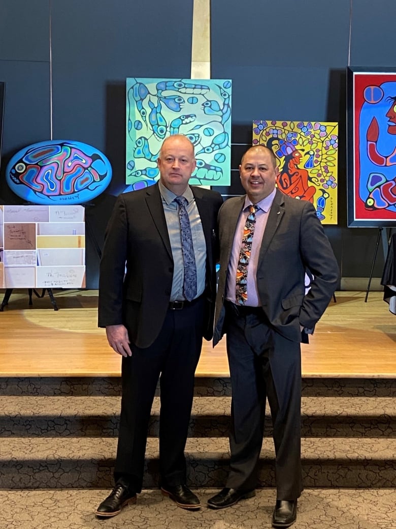 Two men stand in front of a stage containing Indigenous artwork.