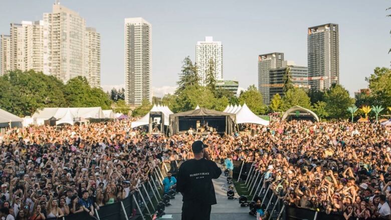 A photo taken from the stage at a music festival shows a musical artist from behind, dressed all in black and holding a microphone. They stand in front of a large crowd of people in a park, with skyscrapers in the background.