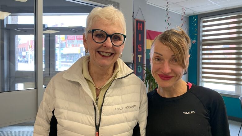 Two women, one older with short white hair wearing a white winter jacket and one younger wearing a black track suit smile against an office background