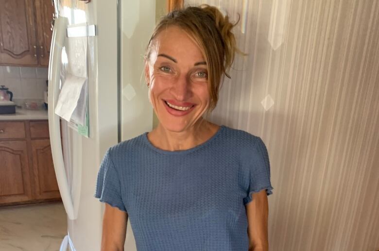 A woman, about forty years old, wearing a blue shirt with blonde hair pinned up smiles widely against the background of a kitchen