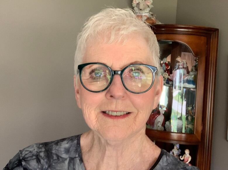 A white-haired woman smiles against the backdrop of a china cabinet in a living room