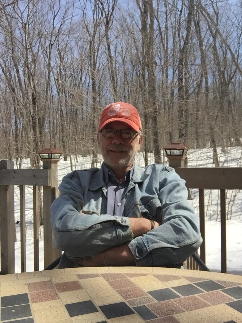 A man wearing a jean jacket and red ball cap sits with arms folded, smiling, against a backdrop of snow and trees.