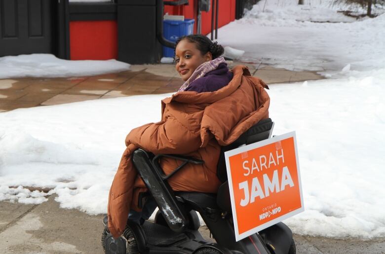 A woman in a wheelchair driving on the sidewalk with an orange campaign sign that says Sarah Jama NDP