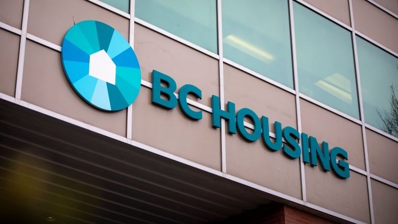 A blue circular symbol, with a house within it, next to a sign that reads 'BC Housing' is affixed on a building.