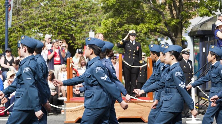 Young military cadets are shown from the side, dressed in blue uniforms, as they march in rows of three during a parade. A saluting captain dressed in black can be seen in the background.