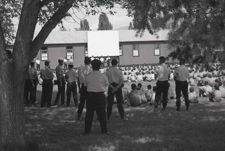 A black and white photo shows men and boys dressed in white shirts and black pants, with military hats, standing beneath a tree with their backs to the camera. In the background, several rows of seated boys can be seen looking at a stage, where four cadets leaders are standing.