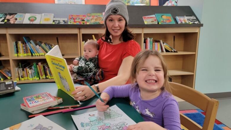 A woman reads a book to her infant son while her daughter colours.