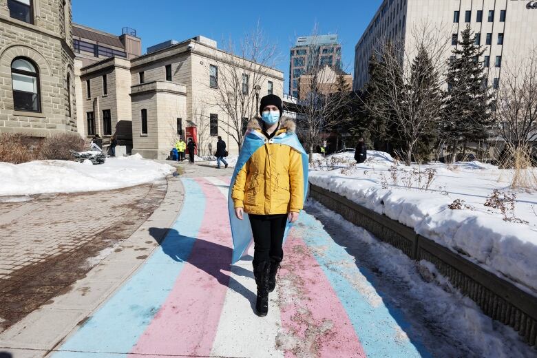 A person with a pink, blue and white flag draped over their shoulders walks across a stretch of sidewalk painted in the same colours.