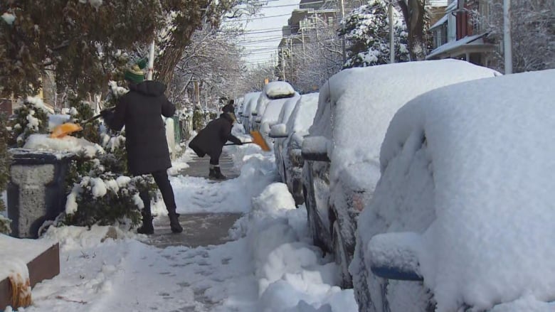 People shovel snow off the sidewalk and cars.