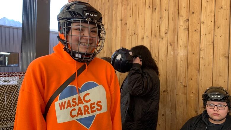 A girl in an orange sweater and a hockey helmet.