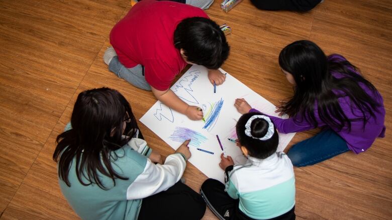 Kids sit on the floor colouring on a big piece of white paper.