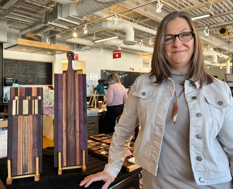 A middle-aged woman smiles for the camera. Next to her on a table are two wooden cutting boards on stands.