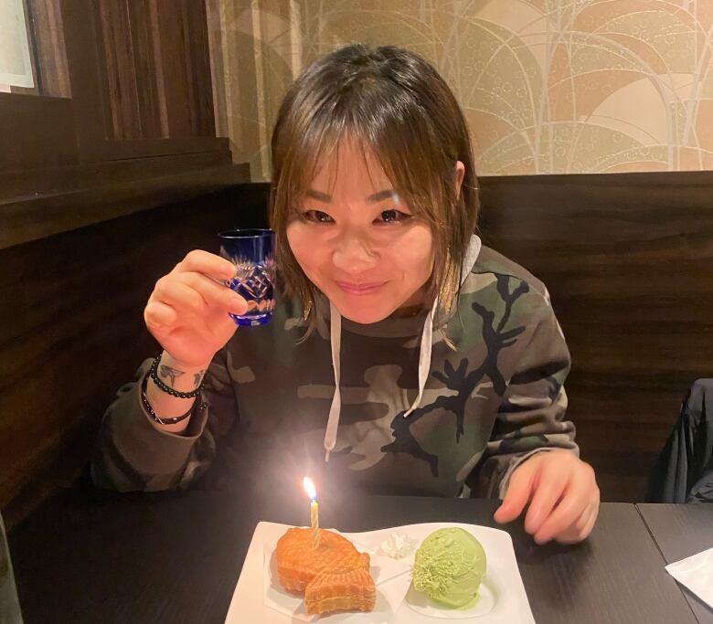 A woman sits on a table and holds a small glass with her right hand, with a plate of dessert and candle in front of her.