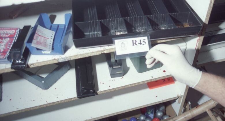 A hand wearing a white glove holds up a tag reading R45 in front of a shelf speckled with blood and a black cash box with change inside 