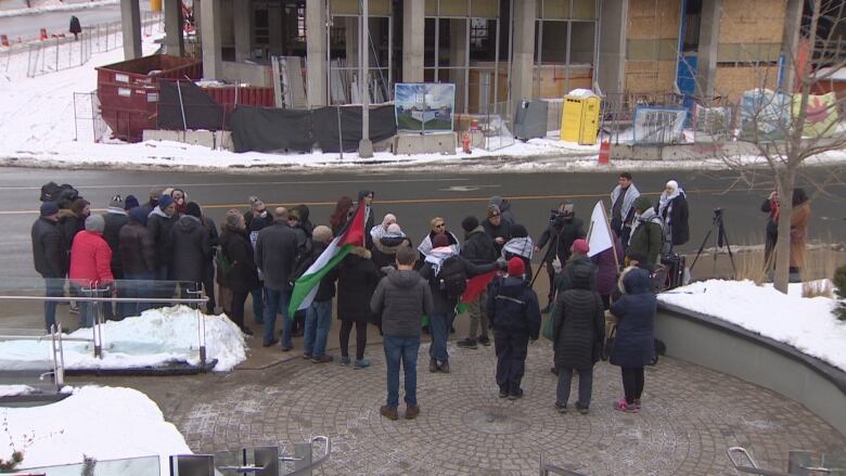 A group of people gather on the sidewalk during a protest, Some are wearing keffiyahs and a person is carrying a Palestinian flag.