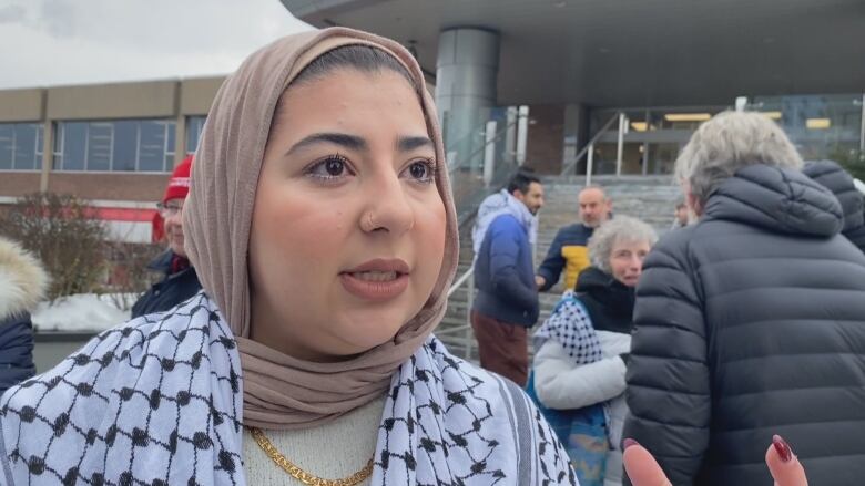 A woman in a hijab and wearing a keffiyeh speaks to the camera.