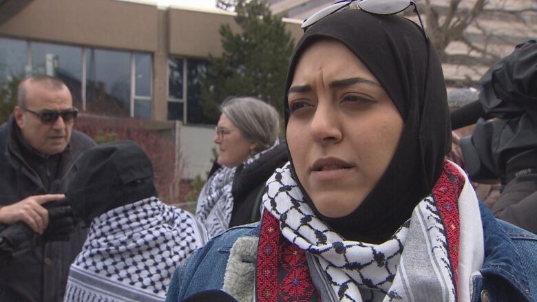 A woman in a denim jacket and a keffiyeh looks at the camera.