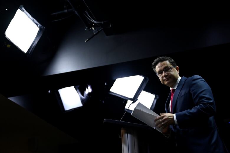 A politician leaves a lectern with his papers as TV lights shine on him.