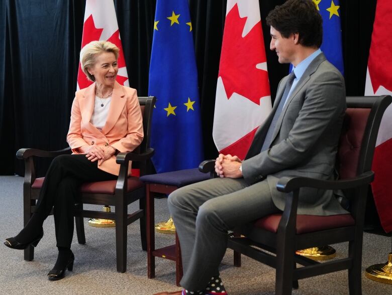 Ursula von der Leyen sits next to Prime Minister Justin Trudeau, both on chairs in front of European Union and Canadian flags.