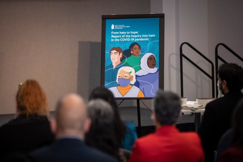 Rows of people sit in chairs in a conference room. A sign with an illustration of people wearing masks is in the background.