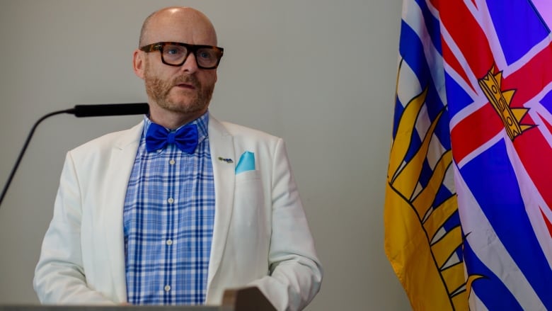 A bald man with glasses and beard wearing a white suit coat and a bow tie speaks from the rostrum with the B.C. provincial flag in the background.