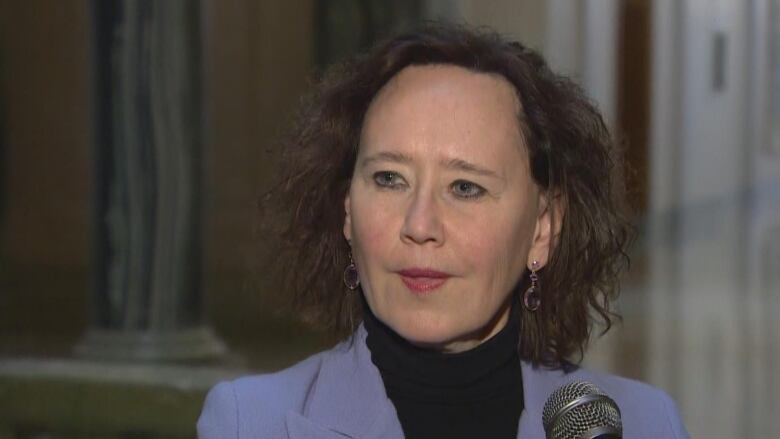 A white woman with short, wavy brown hair stands in front of a microphone. She's wearing a black turtleneck under a lavender blazer. 