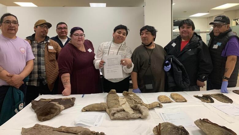 A group of people stand along a table with a number of caribou-skin items, including a baby winter suit. 