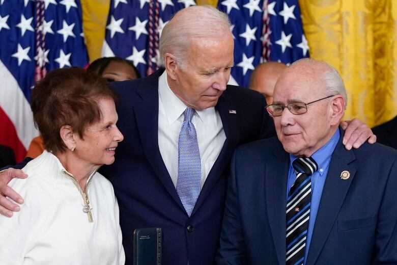 A man in suit puts his arms around a man and a woman on either side.