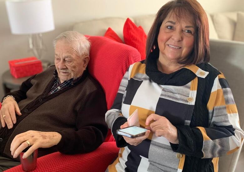 An elderly man sits in a red armchair on a bright, sunny day with a younger woman sitting on another chair to his right, holding a cellphone.