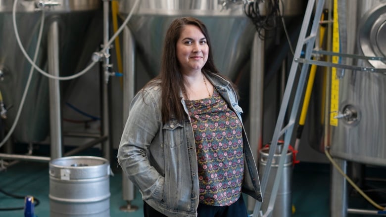 A woman stands inside a brewery. Behind her are  stainless steel fermentors and other equipment. 