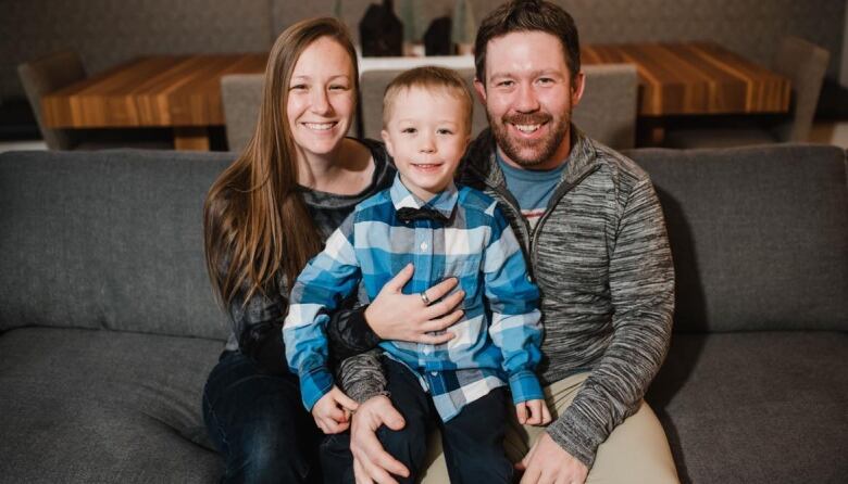 A mom and dad sit on a grey couch in their living room with their young son between them, on their laps.